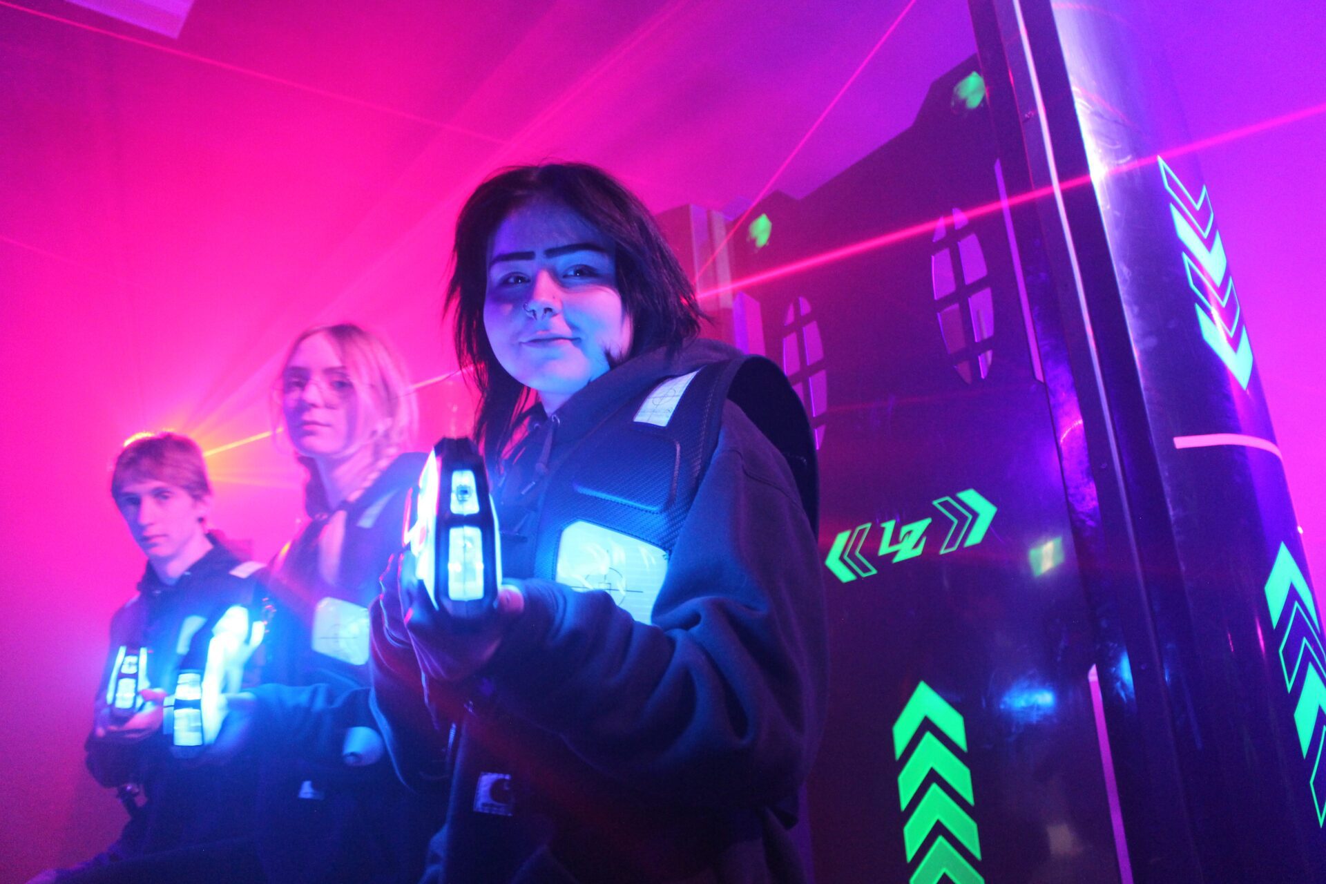 Laser Tag Teens Three teenagers - a boy and two girls - stand in a staggered line next to a black wall with flourescent yellow arrows painted on it. The atmosphere is smokey and there are red laser beams cutting through the background. There are pink, purple, and blue lights colouring the smoke and the faces of the teens. All three hold laser tag guns - about the length of their forearms and glowing with blue light. They point their guns at the camera. The two teens in the background have serious expressions on their faces. The teen in the foreground - a girl - has a small smile on her face as the looks straight into the camera and points her gun.